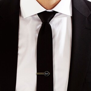 May include: A black tie with a gold tie clip that says "0.6" on it. The tie is worn with a white dress shirt and a black suit jacket.