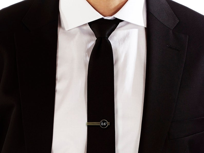 May include: A black tie with a gold tie clip that says "0.6" on it. The tie is worn with a white dress shirt and a black suit jacket.