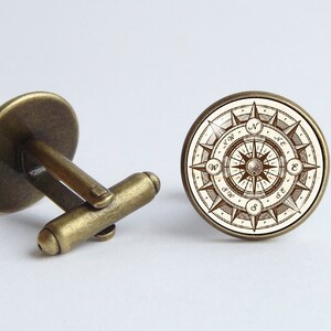 Peut inclure: Une paire de boutons de manchette en or antique avec un motif de boussole. La boussole pr&eacute;sente un fond blanc avec des lignes et du texte bruns, y compris les directions N, S, E et W.