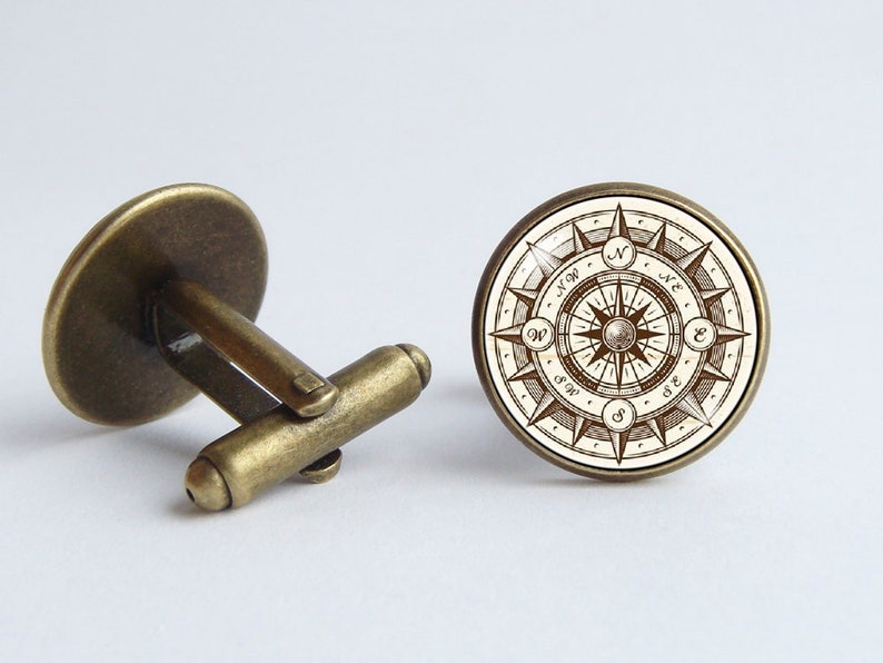Peut inclure: Une paire de boutons de manchette en or antique avec un motif de boussole. La boussole pr&eacute;sente un fond blanc avec des lignes et du texte bruns, y compris les directions N, S, E et W.