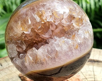 May include: A spherical geode with a polished exterior and a crystalline interior. The outer shell is a mix of brown and gray, while the inside reveals a cluster of light purple and golden crystals. The geode is displayed on a wooden base.