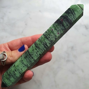 May include: A green and black zoisite crystal tower with small red ruby inclusions. The polished gemstone is rectangular with a pointed top and bottom. The crystal is held in a hand with blue nail polish.