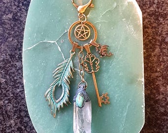 May include: A bronze-colored keychain with a pentagram charm, a feather, a key, and a clear quartz crystal pendant. The keychain is attached to a gold-colored clasp. The background is a green stone.