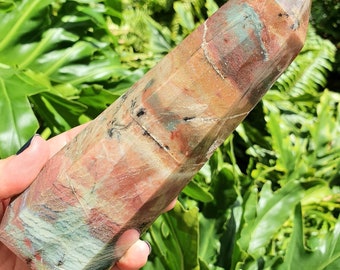 May include: A large, multi-colored, polished, crystal point with a pointed tip. The crystal is a mix of brown, red, green, and white colors. The crystal is being held in a hand.