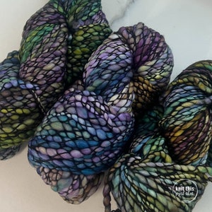 K&ouml;nnte beinhalten: Nahaufnahme von mehreren Str&auml;ngen handgef&auml;rbter Wolle in Lila-, Blau-, Gr&uuml;n- und Gelbt&ouml;nen. Das Garn hat ein dickes, strukturiertes Aussehen mit einem gestrickten oder gewebten Look. Die Farben verschmelzen und erzeugen einen einzigartigen und k&uuml;nstlerischen Effekt. Das Garn ist wahrscheinlich f&uuml;r Strick- oder H&auml;kelprojekte.