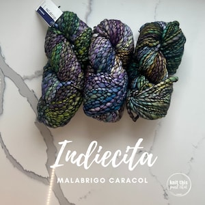 K&ouml;nnte beinhalten: Drei Str&auml;nge Indiecita Malabrigo Caracol Garn in Gr&uuml;n-, Lila- und Blaut&ouml;nen. Das dicke, strukturierte Garn ist auf einer wei&szlig;en Oberfl&auml;che angeordnet. Das Bild enth&auml;lt den Text "Indiecita" und "MALABRIGO CARACOL".