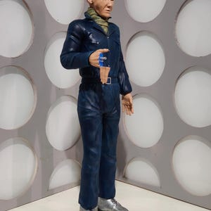 May include: A full-length figure of a bald person wearing a dark blue jumpsuit, silver shoes, and a green and tan scarf. The figure is holding a blue object. The background features a gray wall with white circular accents.