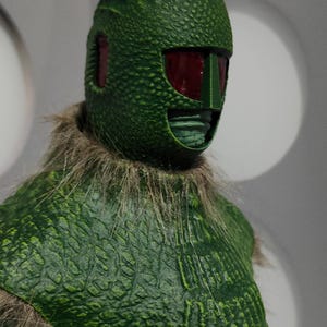 May include: A green, textured figure with a reptilian-like head and red eye coverings. The figure has a textured, scaled appearance and a furry collar. The figure is a collectible item.
