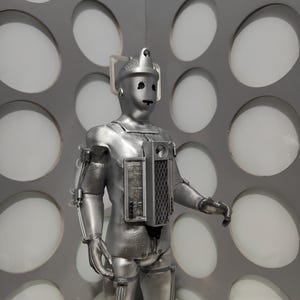 May include: A silver Cyberman action figure with a helmet, chest unit, and articulated limbs. The figure stands in front of a gray wall with large, circular cutouts. The Cyberman has a metallic appearance and detailed mechanical features.