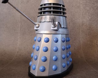 Op de afbeelding: Een zilver en blauw Dalek-model met blauwe bolvormige details. Het model heeft een wapenarm en een zuigervormige arm. De bovenkant heeft een zwart-witte oogsteel en een geel licht. De basis is zwart.