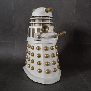 May include: A white and gold Dalek model, a fictional extraterrestrial race from the British science fiction television program Doctor Who. The model features a white body with gold accents, including a gun, eye stalk, and numerous gold spheres.