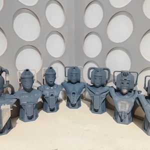 May include: A collection of seven gray Cyberman busts, each with unique helmet and chest plate designs. The busts are arranged in a row, showcasing variations in their robotic features. The background features a wall with large, circular cutouts.