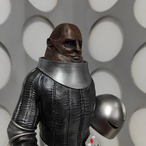 May include: A detailed figurine of a bald, dark-skinned humanoid character with a smooth, rounded head. The figure wears a black, vertically-stitched textured suit with a silver collar, a silver belt, and a small control panel. A silver helmet is held.