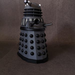 May include: A gray and black Dalek model, a fictional extraterrestrial race from the British science fiction television program Doctor Who. The model features a gun, a manipulator arm, and numerous black spheres. The Dalek has a cylindrical body and a sloped base.