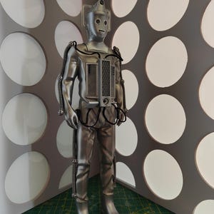 May include: A silver-colored Cyberman action figure, standing upright. The figure has a cylindrical head, a rectangular chest piece, and articulated limbs. Black tubing and wires are attached to the figure's body. The background features a gray wall with white circular cutouts.