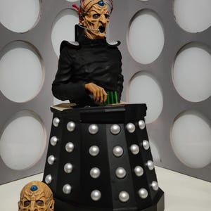 May include: A detailed sculpture of a character from a science fiction series. The figure has a pale face, black attire, and a headpiece. The sculpture is mounted on a black base with silver studs, and a separate head is displayed nearby. The background features a wall with white circles.
