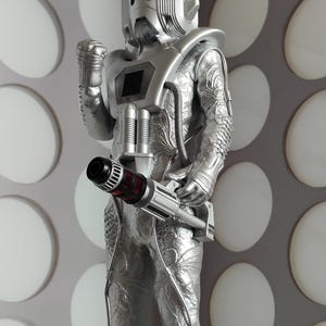 May include: A silver Cyberman action figure, a fictional character from the Doctor Who series, stands with a weapon. The figure has a detailed metallic design, including a helmet, chest unit, and textured suit. The weapon has a red and black barrel.