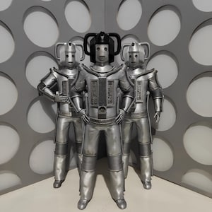 May include: Three silver Cybermen figures stand in a row against a gray wall with circular cutouts. The Cybermen have cylindrical helmets, chest plates, and segmented limbs. The central figure has a distinctive headpiece. The figures are posed with their hands on their hips.