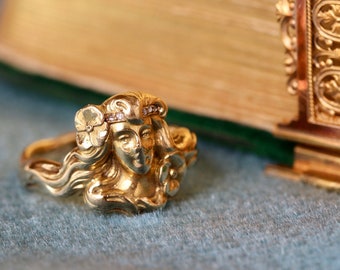 May include: A gold ring with a detailed design of a woman's face with flowers in her hair. The ring is set with small diamonds.
