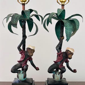 May include: Pair of vintage table lamps featuring monkey figures climbing palm tree-shaped supports. The monkeys wear red jackets and white hats, perched on a blue-green sphere. The lamps have green palm fronds and sit on black square bases.