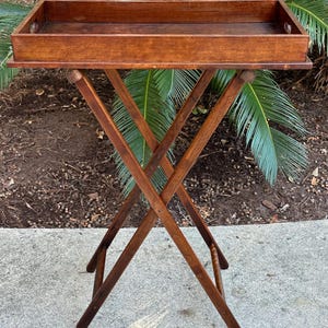 May include: A vintage wooden serving tray table with a rectangular tray and folding X-shaped legs. The tray is a rich brown color, and the legs are a darker shade of brown. The table is set outdoors, with greenery in the background.