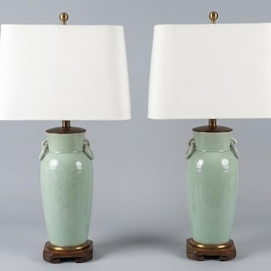 May include: A pair of table lamps featuring pale green ceramic vase-shaped bases with ring handles, dark wood circular bases, and rectangular white fabric shades topped with small brass finials. The ceramic bodies display subtle embossed patterns.