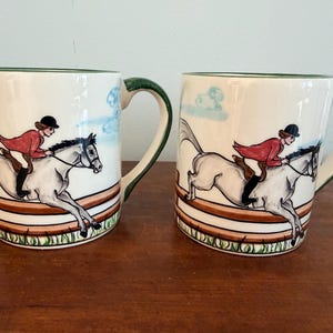 May include: Two ceramic mugs with a horse-riding theme. Each mug features a white background with a hand-painted illustration of a rider in a red jacket on a gray horse jumping over a fence. The rims and handles are green.