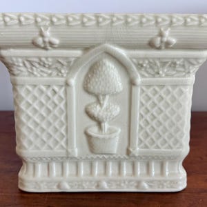 May include: Off-white rectangular ceramic planter with a detailed relief design. The planter features a central arched panel with a potted topiary, surrounded by lattice and floral patterns. Decorative bee motifs adorn the top rim.