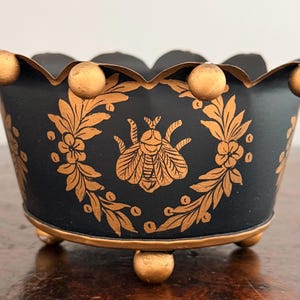 Puede incluir: Un cuenco de metal negro con un borde festoneado que presenta detalles esféricos de color dorado. El exterior está decorado con un gráfico de insecto dorado centrado dentro de una corona floral. El cuenco se apoya sobre tres pequeñas patas esféricas doradas.