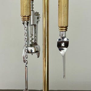 Op de afbeelding: Een goudkleurige bar-gereedschapsset met een zwarte basis. De set bevat een kurkentrekker, een lepel en een flesopener. De gereedschappen zijn bevestigd aan een centrale standaard met een decoratief ontwerp.