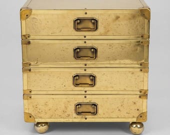 May include: A brass chest of drawers featuring four horizontal drawers, each equipped with a rectangular recessed handle. The exterior is reinforced with metal corner brackets and rivets, and the unit is supported by four spherical feet.