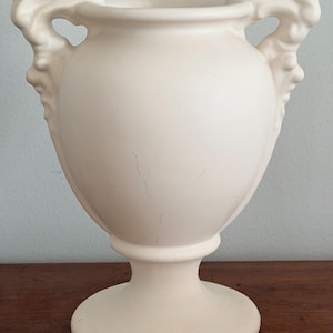 May include: Cream-colored vase with a classic urn shape. The vase features decorative handles with sculpted faces and a pedestal base. The smooth surface has subtle variations in tone, adding to its vintage appeal. A decorative piece for home decor.