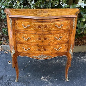 May include: A three-drawer chest with intricate marquetry details and brass hardware. The chest has a curved front and cabriole legs.