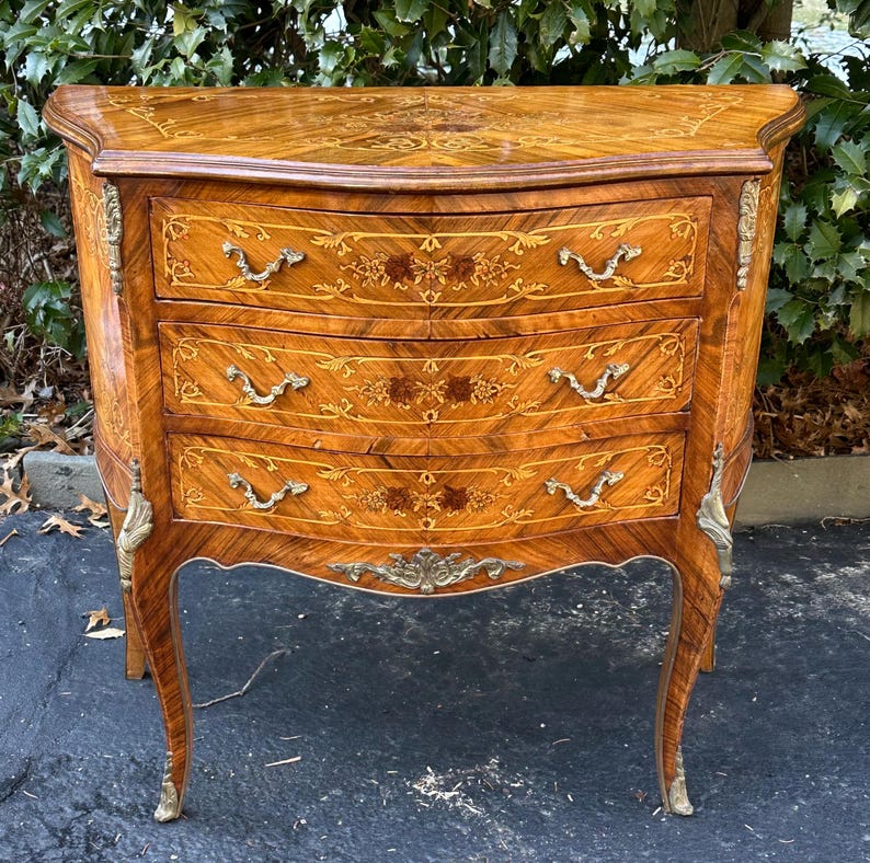 May include: A three-drawer chest with intricate marquetry details and brass hardware. The chest has a curved front and cabriole legs.