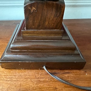 May include: A dark brown wooden lamp base with a square base and a round top. The base has a carved design and a cord coming out of the bottom.