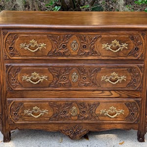 Puede incluir: Cómoda de madera de tres cajones con intrincadas tallas florales y herrajes de latón. La cómoda es de color marrón cálido, con tallas detalladas alrededor de los cajones y en los laterales. Los tiradores y cerraduras de latón añaden un toque de elegancia.