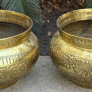 May include: Two large, ornate, gold-colored metal planters with intricate floral designs. The planters are round and have a wide, open top.