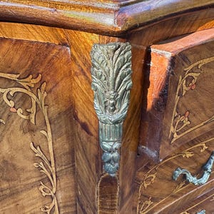 May include: A wooden chest of drawers with intricate marquetry inlay and ornate silver-toned hardware. The chest has a rich brown finish and features a floral design.