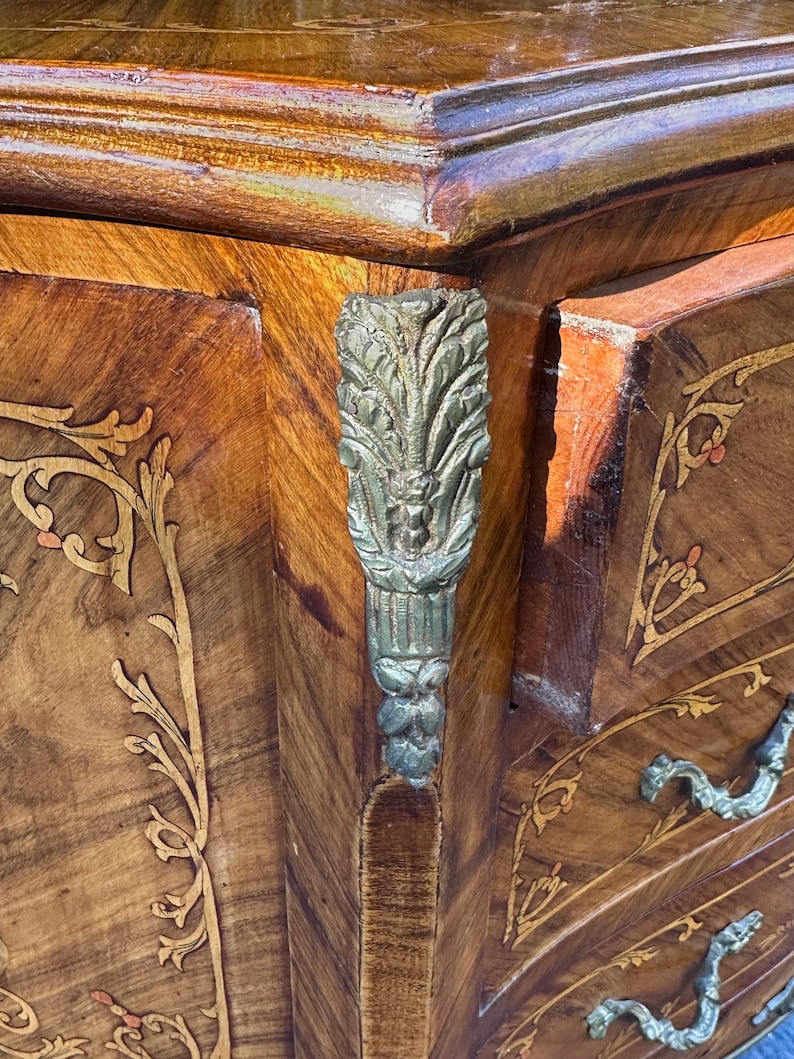 May include: A wooden chest of drawers with intricate marquetry inlay and ornate silver-toned hardware. The chest has a rich brown finish and features a floral design.