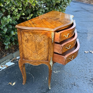 May include: A curved, wooden chest of drawers with three drawers. The chest is made of a light brown wood with a marquetry design. The drawers have brass handles and are open, revealing the interior.