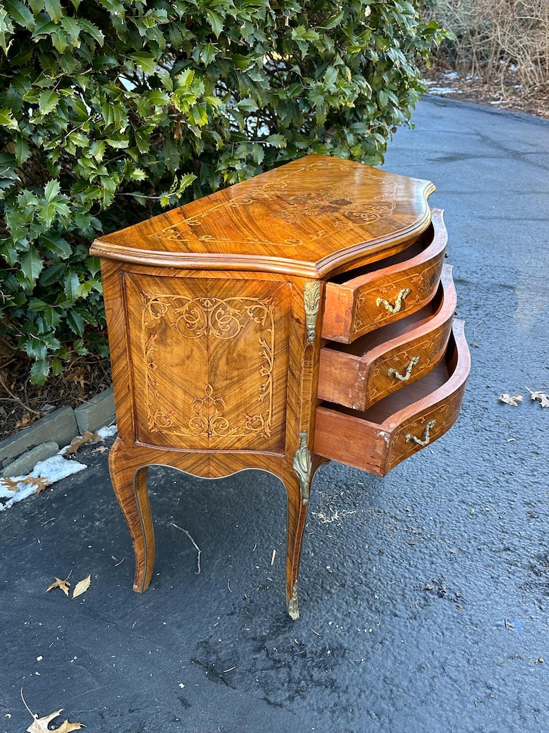 May include: A curved, wooden chest of drawers with three drawers. The chest is made of a light brown wood with a marquetry design. The drawers have brass handles and are open, revealing the interior.