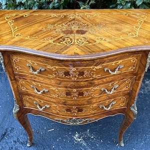 May include: A three-drawer wooden chest with intricate marquetry inlay and brass hardware. The chest has a curved front and sits on cabriole legs.