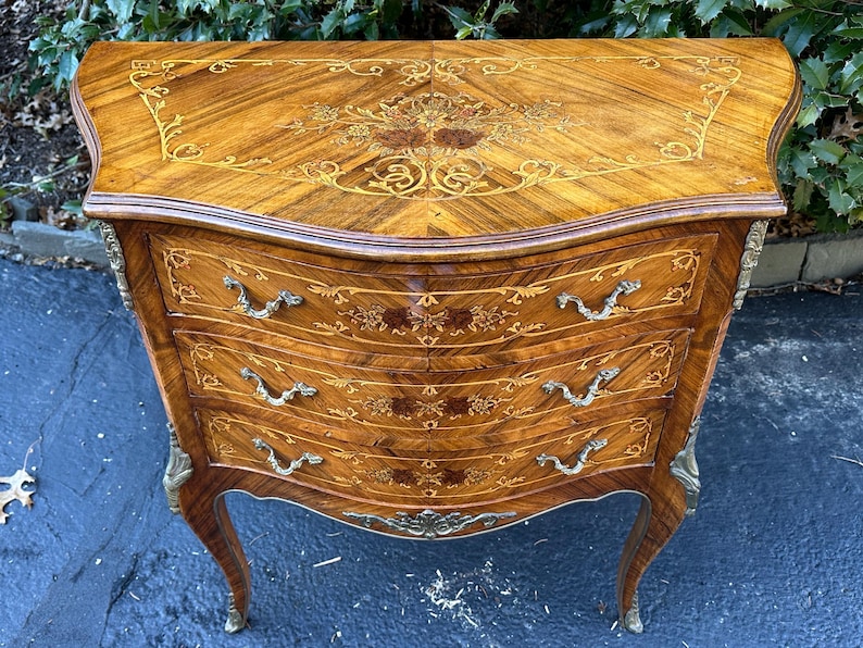 May include: A three-drawer wooden chest with intricate marquetry inlay and brass hardware. The chest has a curved front and sits on cabriole legs.