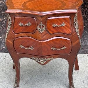 May include: A small, antique wooden chest of drawers with a curved front and legs. The chest is a warm brown color with a patterned wood grain. It features brass hardware, including drawer pulls and decorative accents.