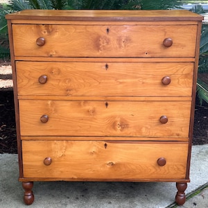 Puede incluir: Un mueble de madera de cuatro cajones con un acabado marrón claro y cálido. Cada cajón tiene un pomo redondo de color marrón oscuro y una cerradura. El mueble tiene un borde marrón oscuro y se apoya en cuatro patas cortas torneadas.