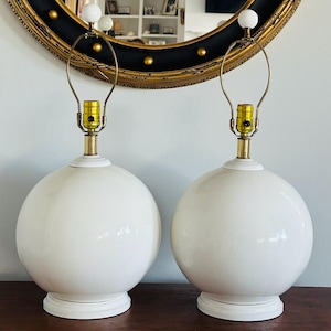 May include: Pair of white ceramic globe table lamps. Each lamp has a round base, a spherical body, and a brass-colored neck supporting a light fixture. The lamps are topped with a white finial and are set against a wall with a decorative mirror.
