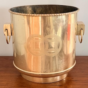 May include: A gold-colored metal ice bucket with a circular base and handles. The bucket features an engraved design of two overlapping circles with a square pattern in the center of each. The handles are square with a ring attached.