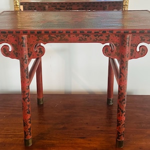 Puede incluir: Pequeña mesa roja con adornos oscuros. La mesa tiene una parte superior rectangular y cuatro patas con adornos decorativos. Las patas están adornadas con patrones florales negros y puntas doradas. Probablemente sea una pieza decorativa.