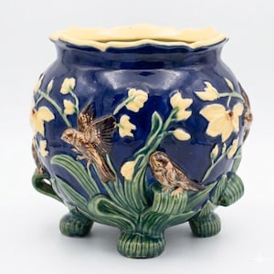 May include: A deep blue ceramic planter featuring raised relief designs of brown birds perched among green leaves and pale yellow flowers. The vessel has a scalloped rim and stands on three small, textured feet, showcasing a glossy finish.