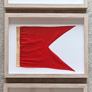 May include: Three framed nautical signal flags. The top flag has blue, red, and off-white horizontal stripes. The middle flag is red with a white triangle. The bottom flag has red, off-white, and blue vertical stripes.